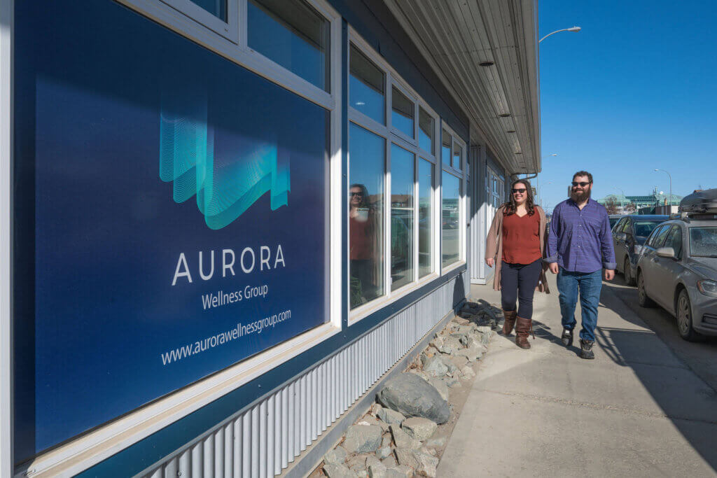 Two people walk on a sidewalk past the Aurora Wellness Group building, featuring large blue signage in the window dedicated to Stephanie Leithead.
