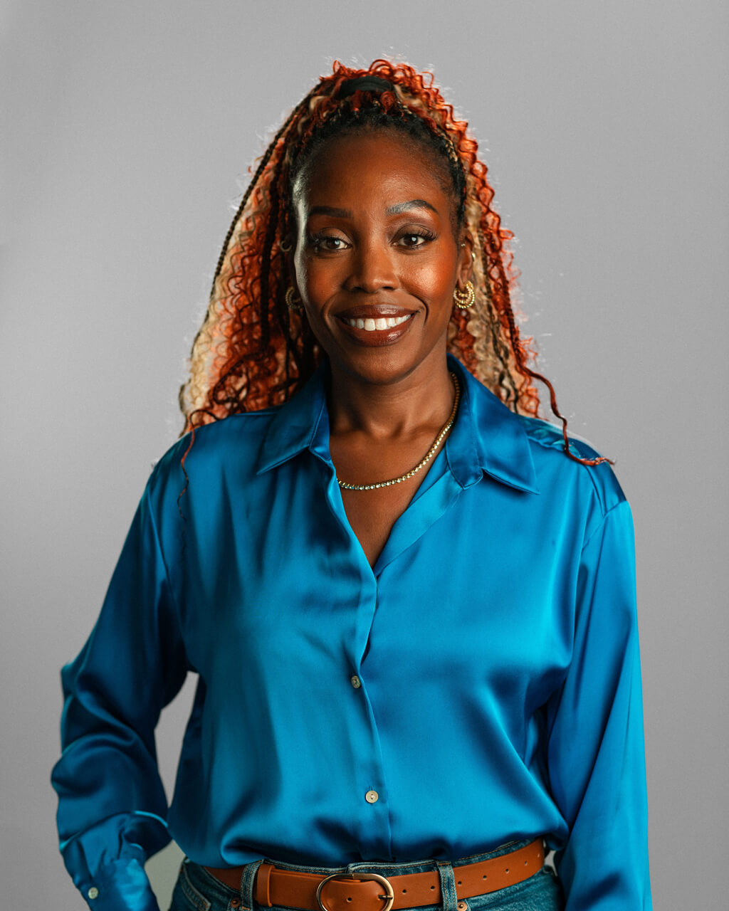 Cadmona Hall, a woman with long curly hair, wears a bright blue satin blouse and gold jewelry as she stands against a plain gray background, smiling at the camera.