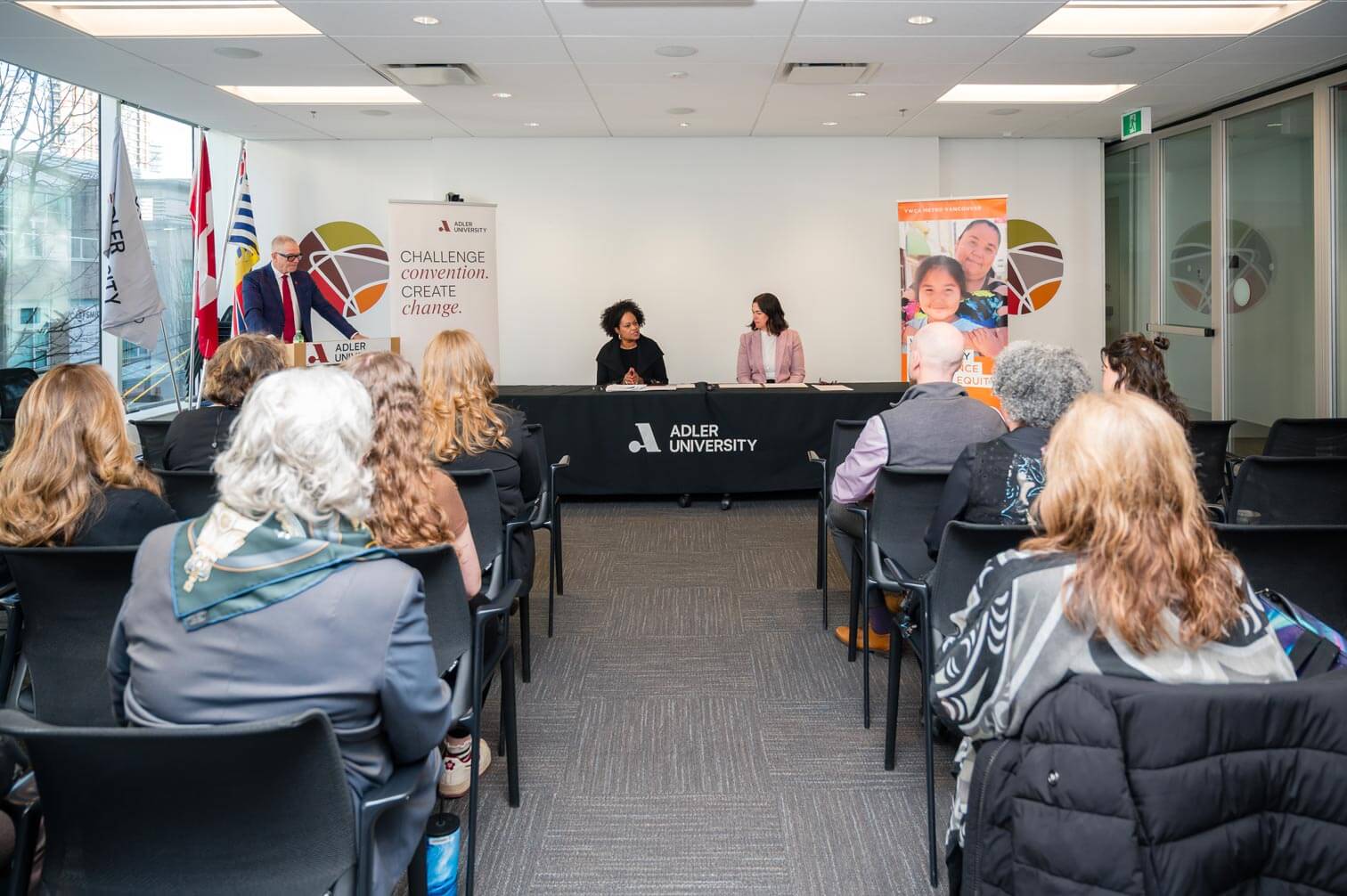 A panel discussion on the behavioural health workforce takes place at Adler University, featuring three speakers at a table and an attentive audience. YWCA Metro Vancouver banners and flags are displayed in the background.