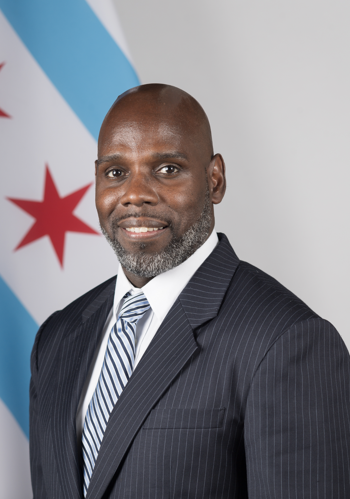 A man in a dark pinstripe suit and striped tie stands in front of a white background with a partial view of the Chicago city flag, representing Joseph Mapp's commitment to education and the Chicago reentry strategy.