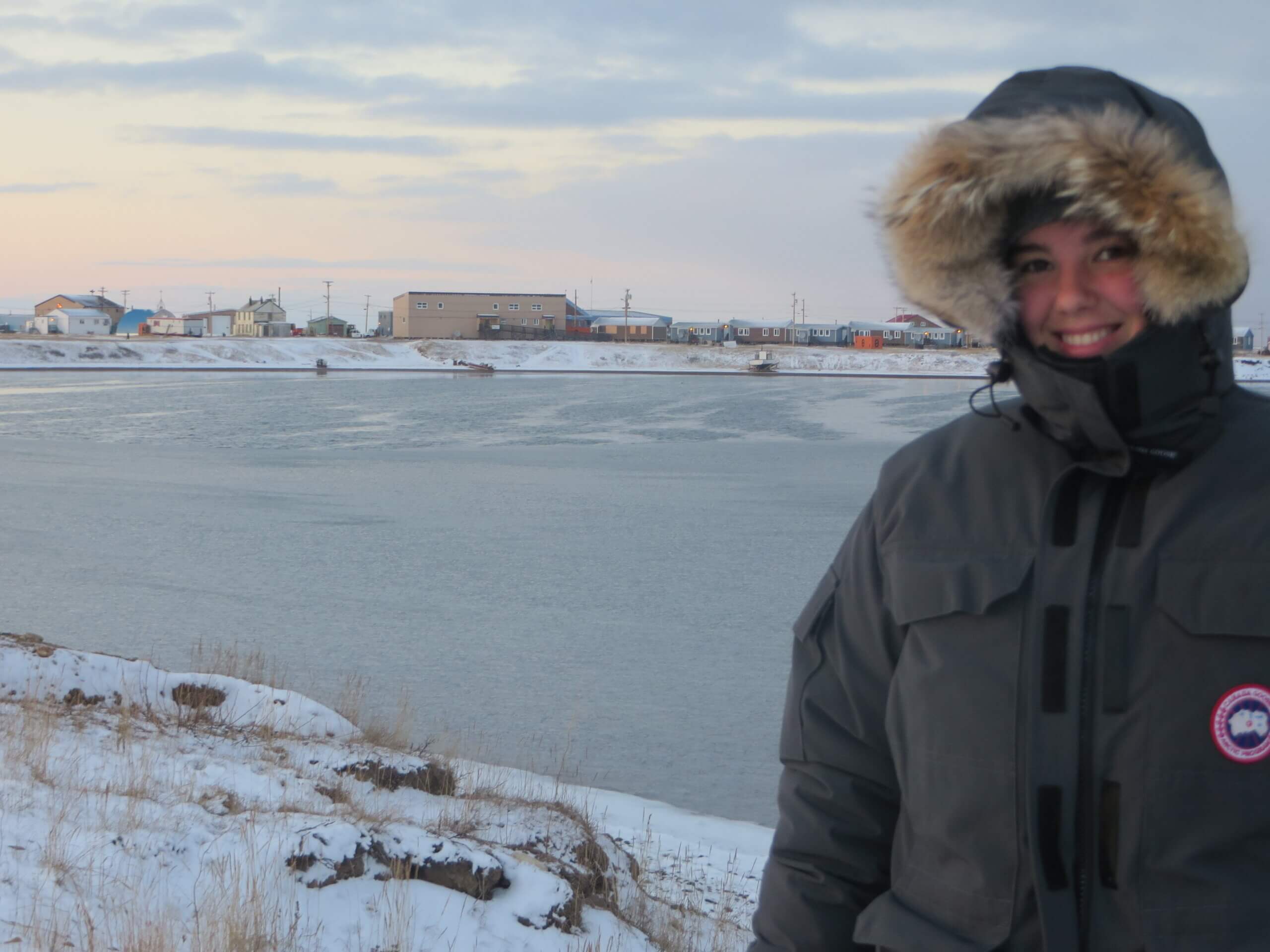 Person in a fur-lined parka stands near a snowy riverbank with houses and buildings visible across the water under a cloudy sky, reminiscent of scenes captured by Stephanie Leithead in her explorations near an Arctic clinic.
