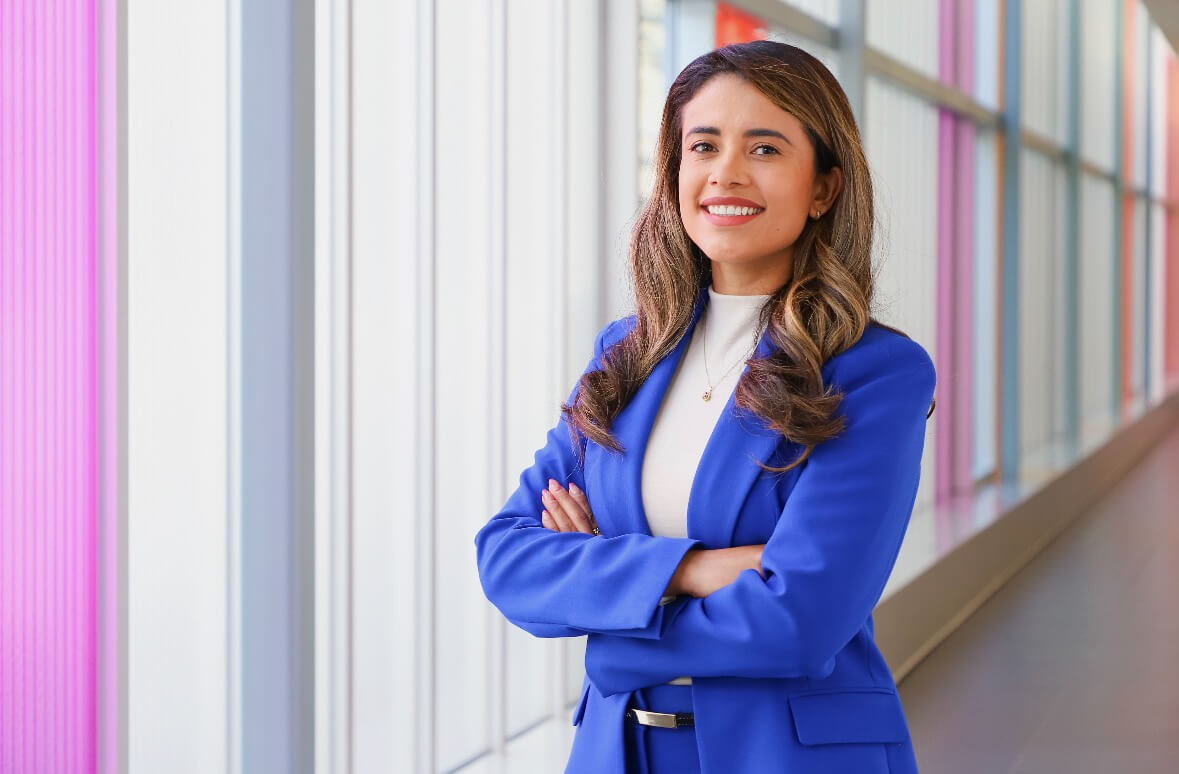 Woman in a blue suit stands with arms crossed, smiling, in a bright hallway with large windows and pink vertical panels.