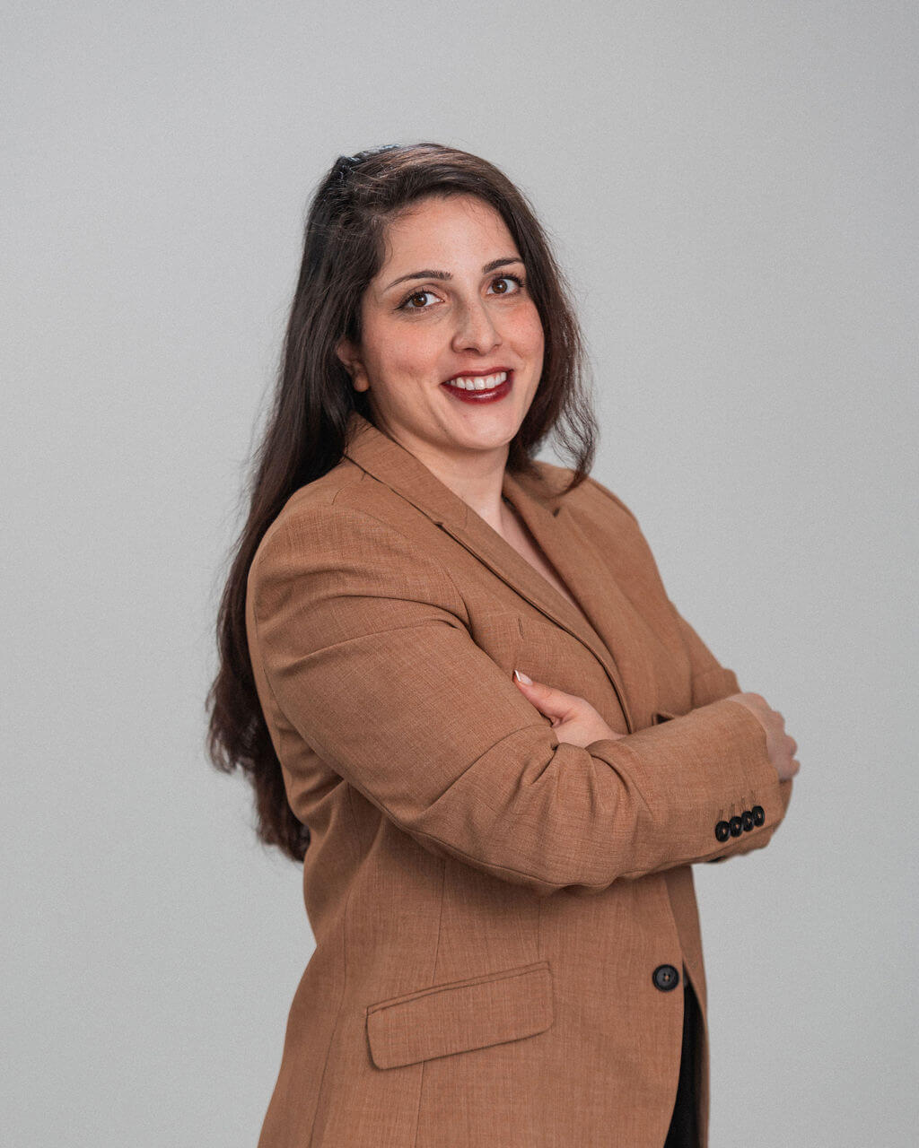 Sara Saeedi, a woman with long dark hair, stands against a plain background in a brown blazer, smiling confidently with her arms crossed.