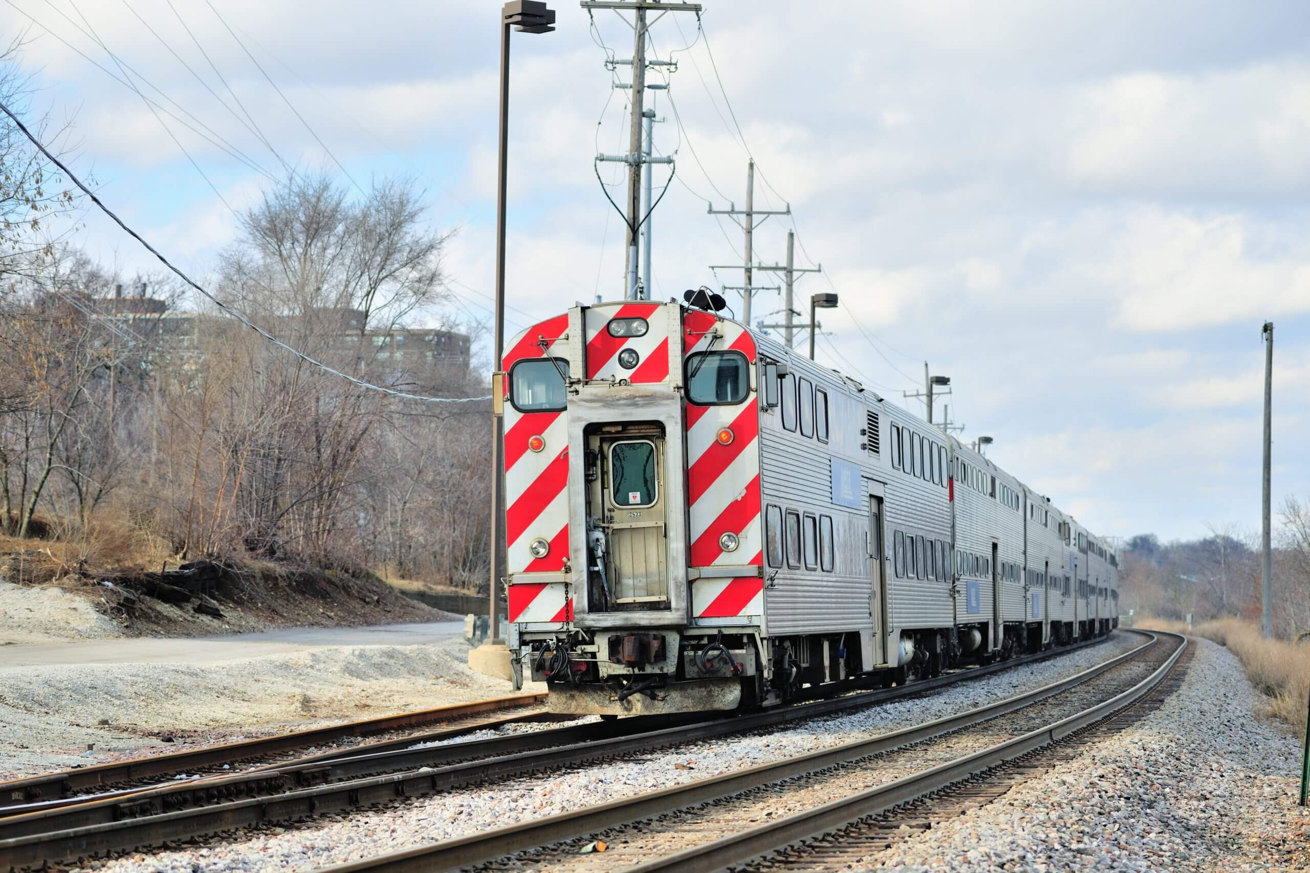 A silver double-decker passenger train with red and white stripes on its rear, possibly moving to Chicago, travels on a curved track beside leafless trees and power lines.