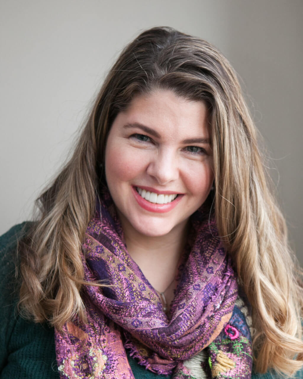 Jennifer Kotecki, a woman with long, light brown hair, is smiling. She is wearing a green sweater and a purple patterned scarf. The background is neutral.