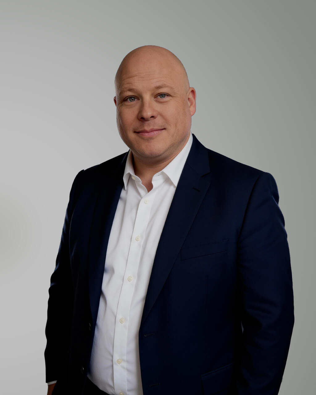 A bald man wearing a dark blue suit and white shirt stands against a plain, light gray background, looking at the camera with a slight smile.