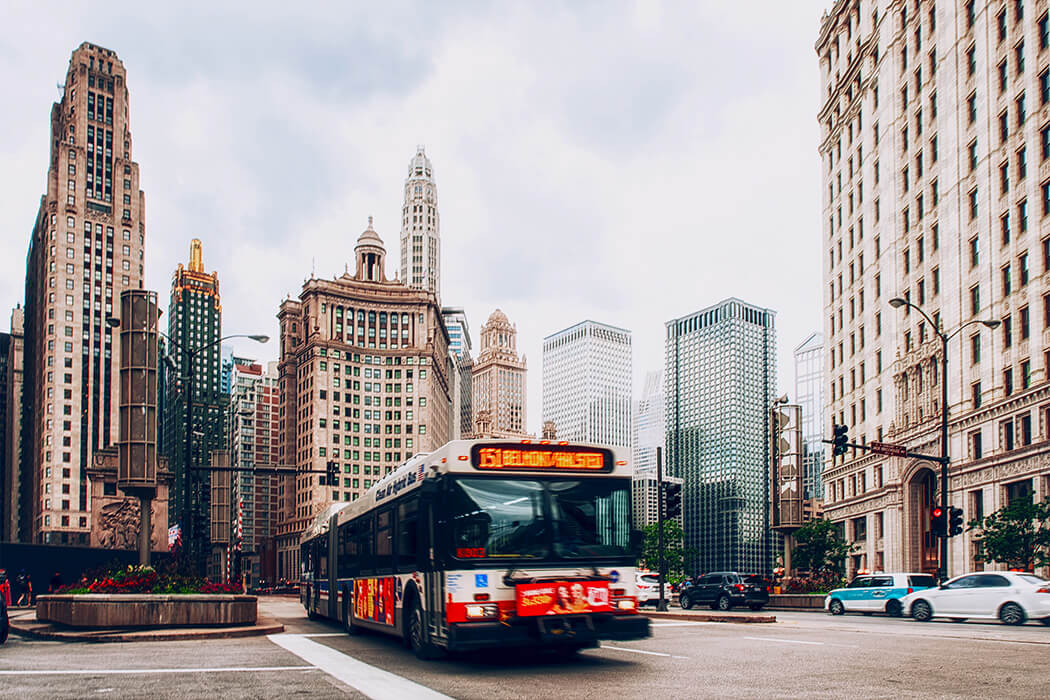 A city bus drives along a downtown street surrounded by tall skyscrapers and historic buildings, capturing the bustling energy that welcomes those relocating to Chicago in this vibrant urban setting.