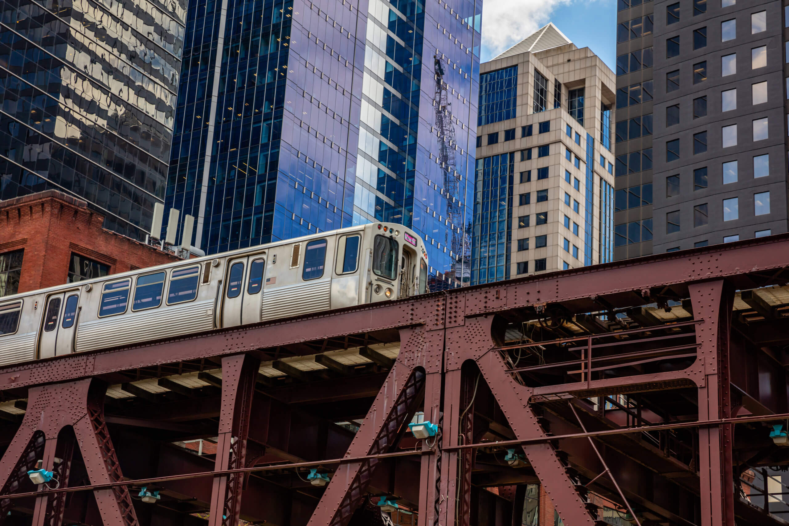 A silver commuter train travels on an elevated steel track in front of tall, modern office buildings—a familiar sight for anyone moving to Chicago or considering a Chicago relocation.