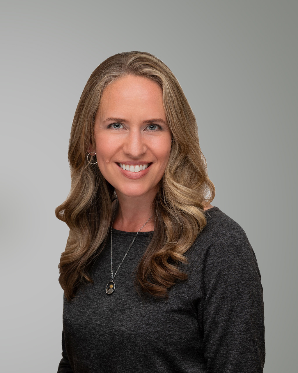 Janelle Kwee, a woman with long, wavy blonde hair wearing a dark Grey top and a necklace, smiles at the camera against a plain Grey background.