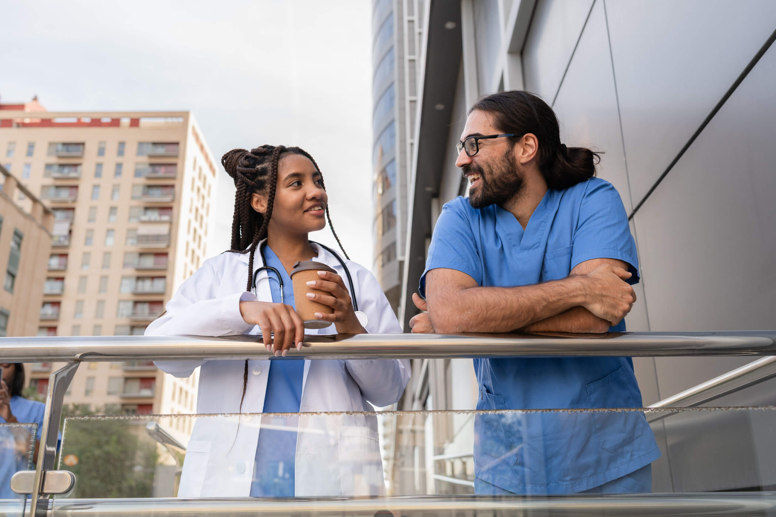 Two healthcare professionals in scrubs and a lab coat stand outside a building, talking and smiling—with one holding a coffee cup—while discussing relocating to Chicago for new career opportunities.