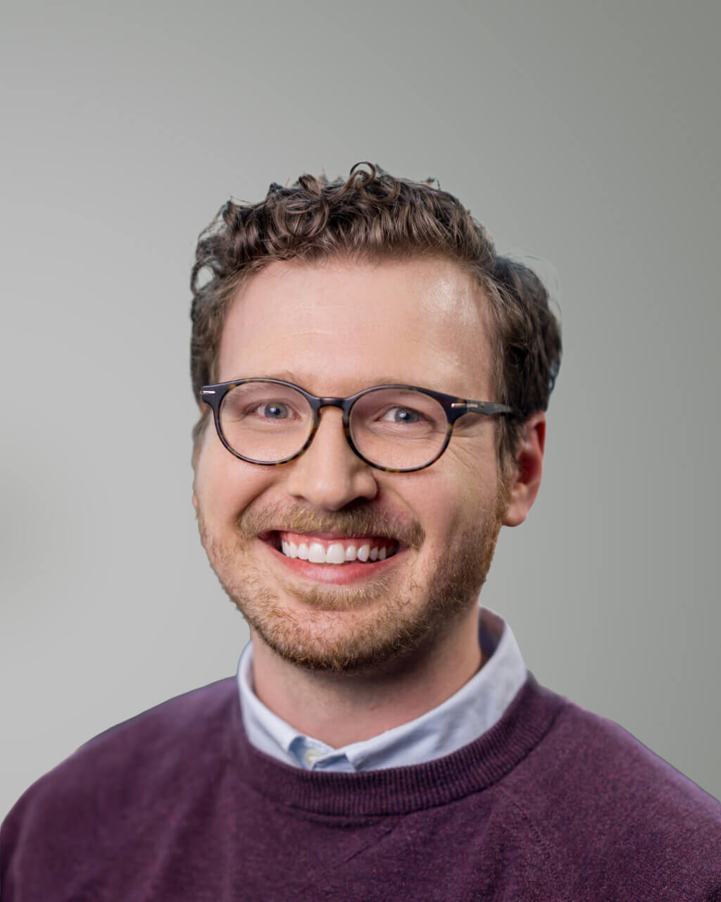 Vincent Marasco, a man with short curly brown hair, glasses, and a beard, smiles at the camera. He is wearing a maroon sweater over a light blue collared shirt. The background is plain gray.