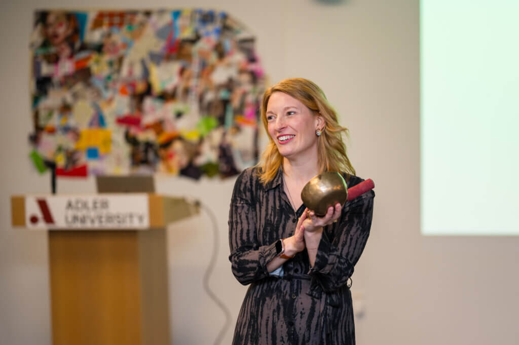 A woman stands indoors in Vancouver, smiling as she holds a sculptural object, with an Adler University podium and a colorful collage in the background—capturing the creative spirit of art therapy.
