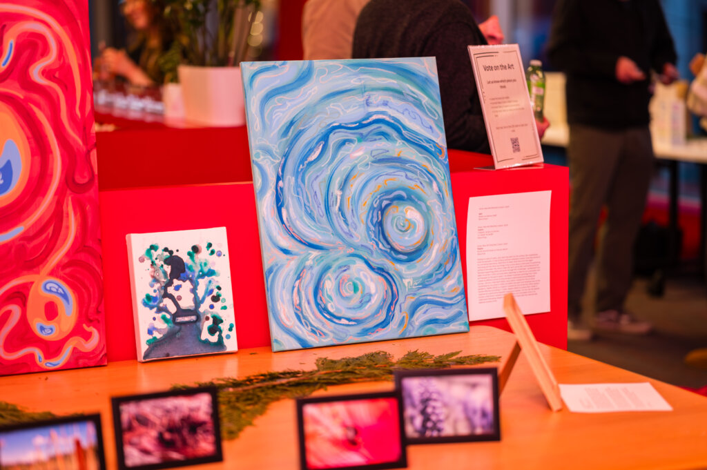 A table displays several pieces of art, including a large blue abstract painting, a smaller green and blue painting, and framed photographs, at an indoor Adler University exhibit in Vancouver focused on art therapy.