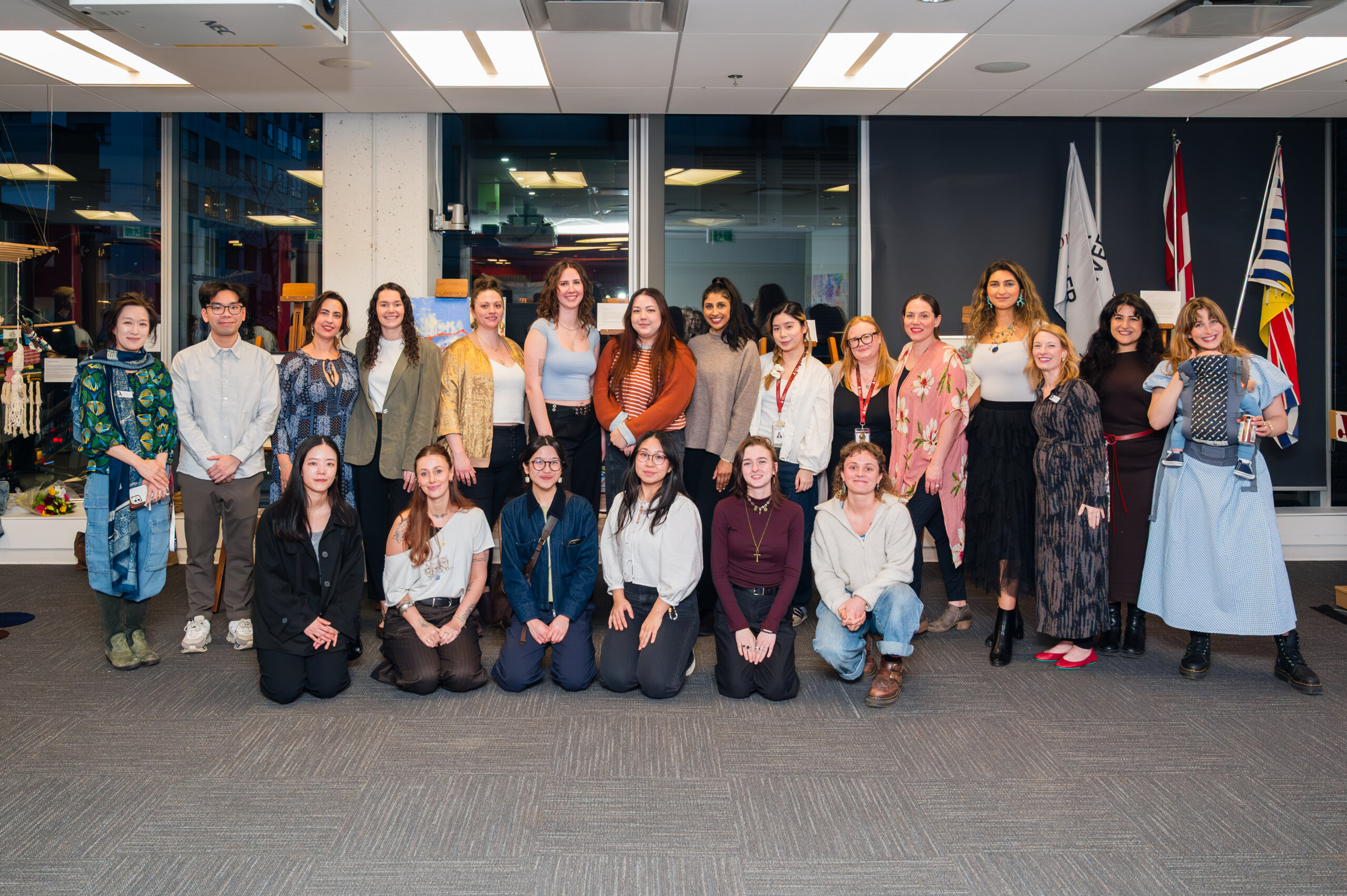 A group of 22 people pose for a photo indoors at Adler University, with some standing and others kneeling. Office windows and flags are visible in the background, capturing the spirit of an art therapy event.