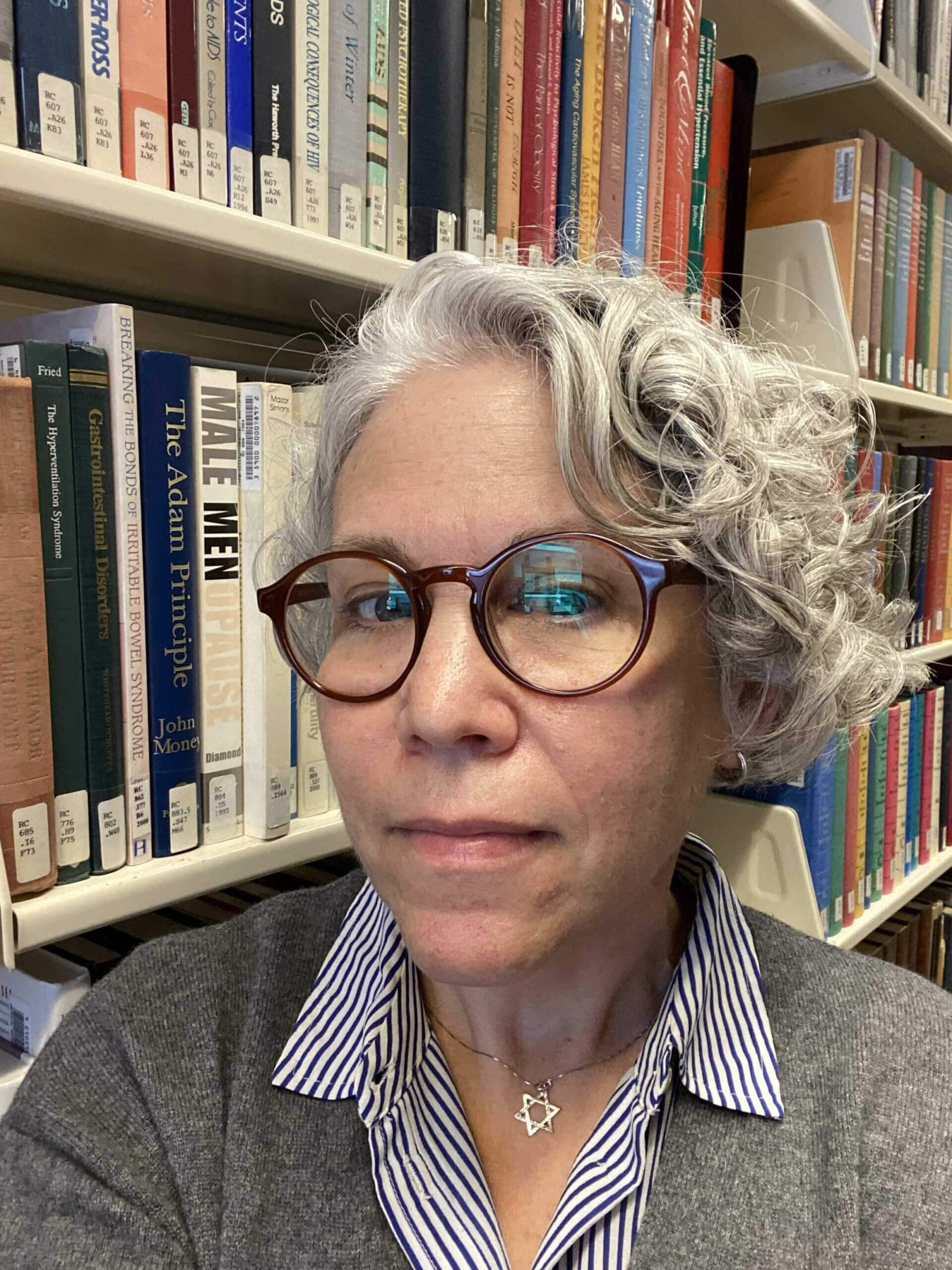 Ariel Orlov, with short gray curly hair and glasses, stands in front of bookshelves in a library.