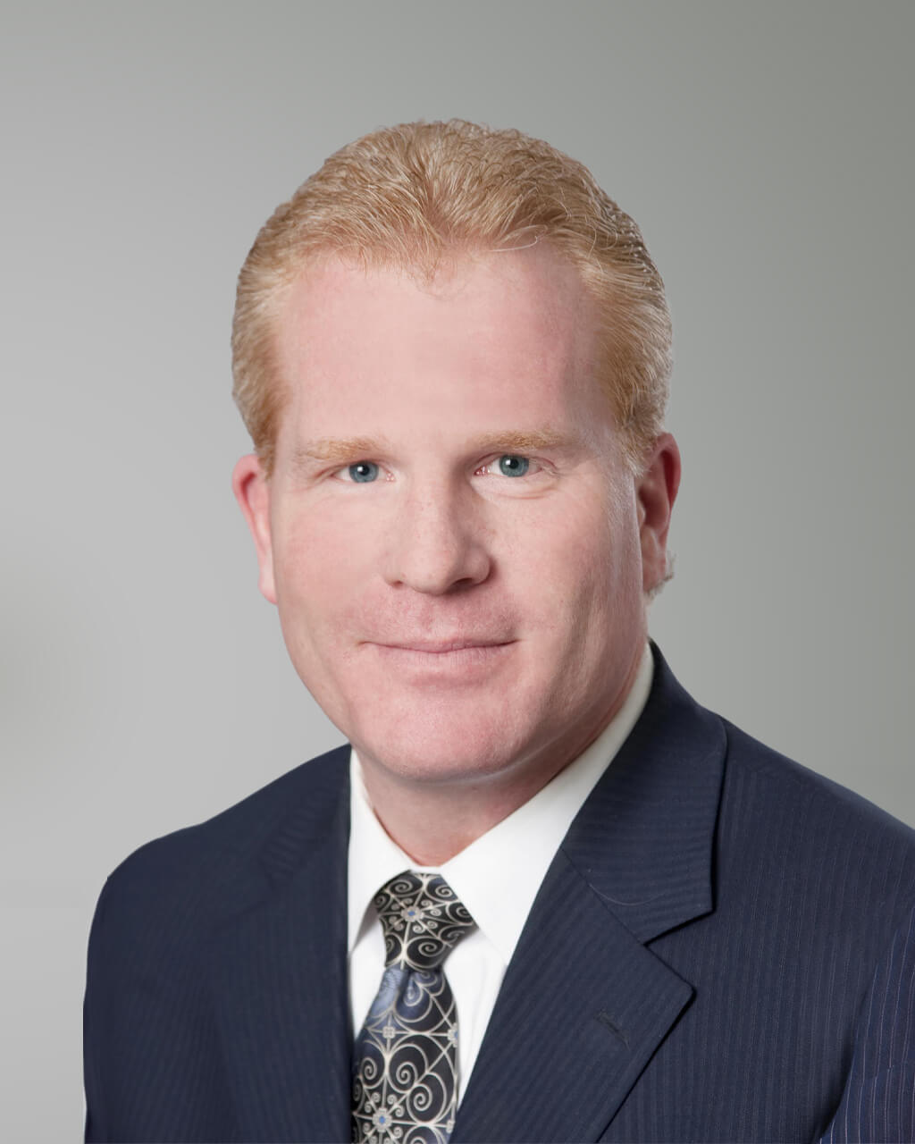 A man with short, light reddish-blonde hair, identified as Deane M. Rabe, wears a dark suit, white shirt, and patterned tie while posing against a plain light gray background.
