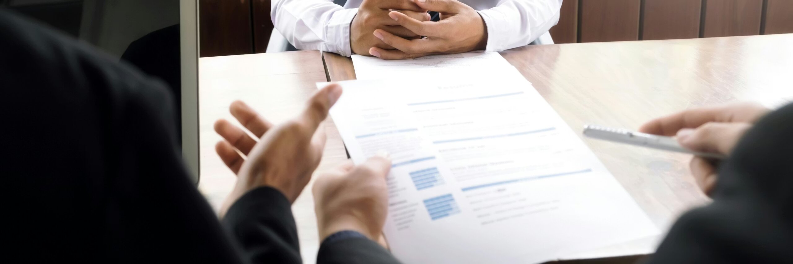 Two people sit across a desk; one, likely from HR, holds a resume and gestures, while the other—possibly among early-career workers—sits with hands clasped, suggesting a job interview setting.