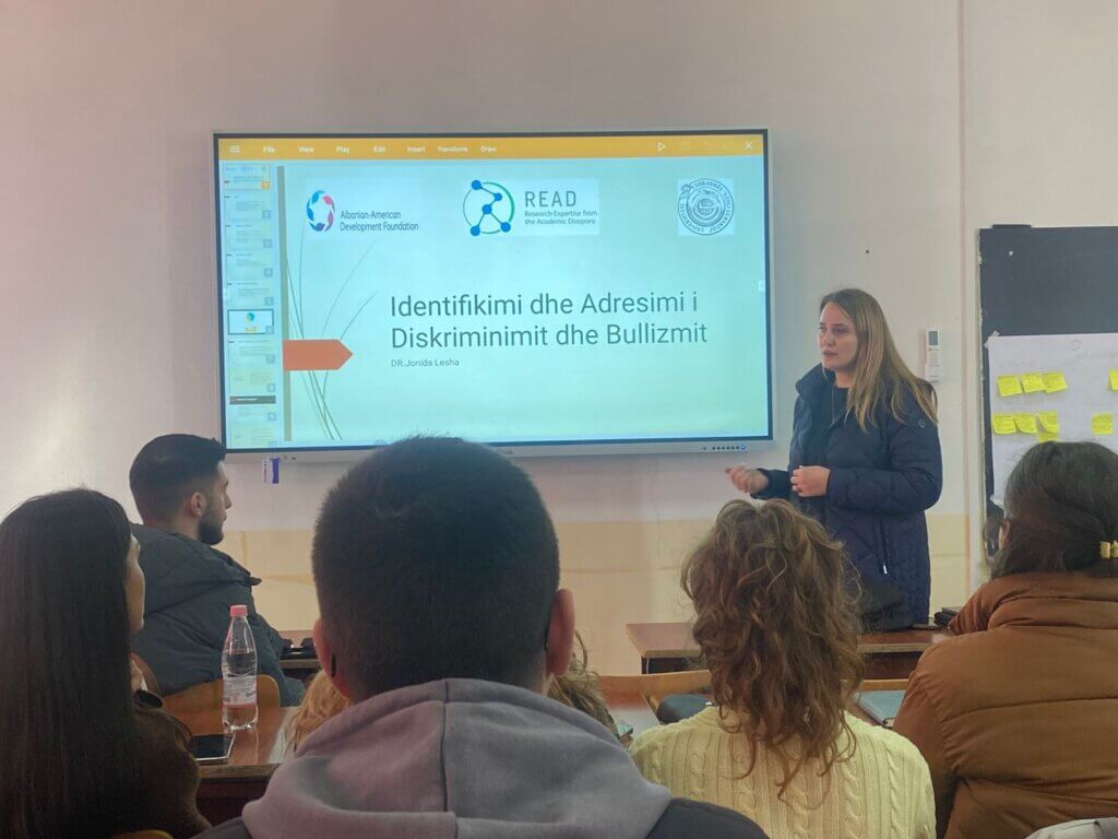 Elvita Kondili stands in front of a classroom at an Albanian university, giving a presentation titled “Identifikimi dhe Adresimi i Diskriminimit dhe Bullizmit” to a seated audience.