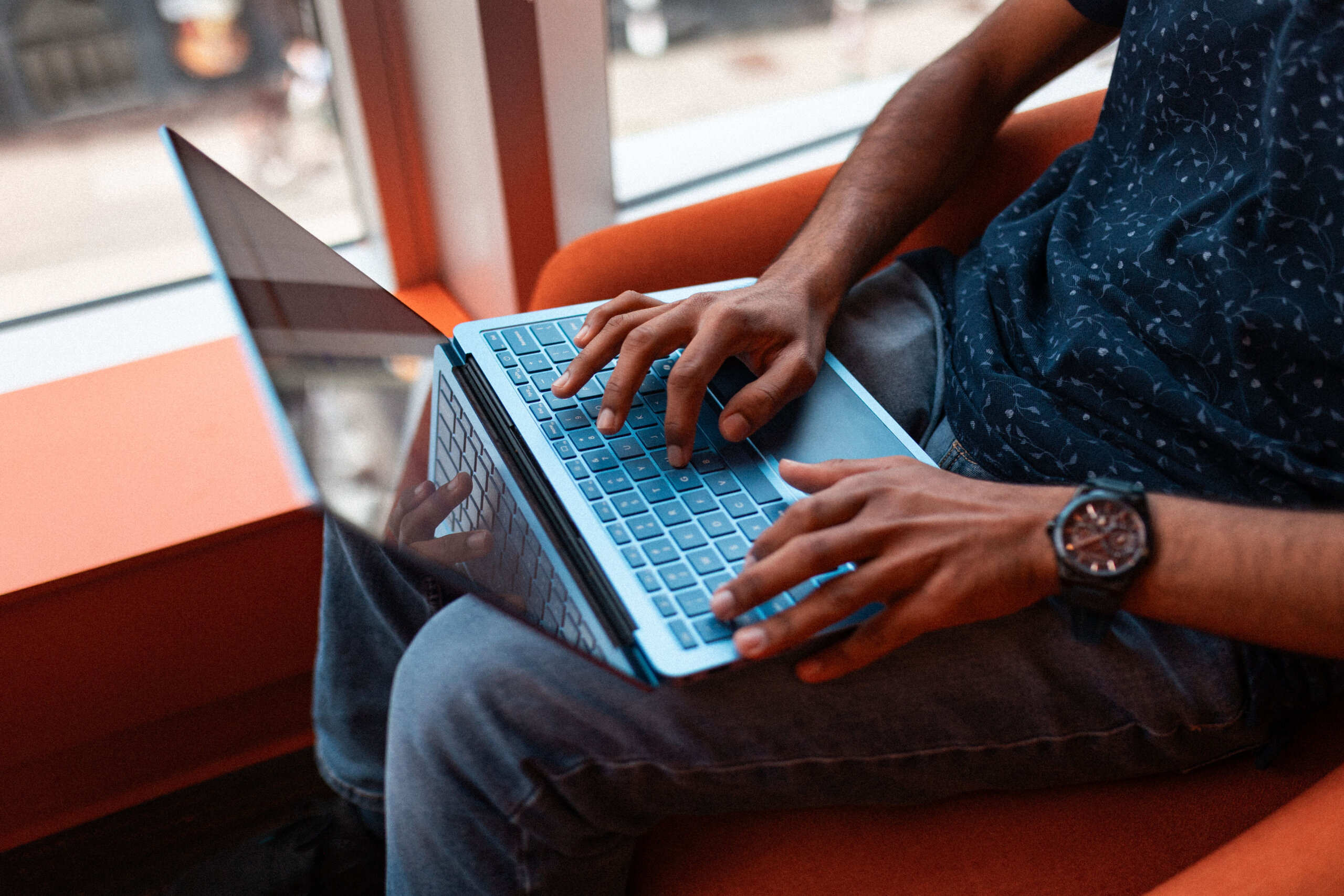 A person sits on an orange chair by a window, typing on a blue laptop placed on their lap, researching Devant opportunities through the career resource center.