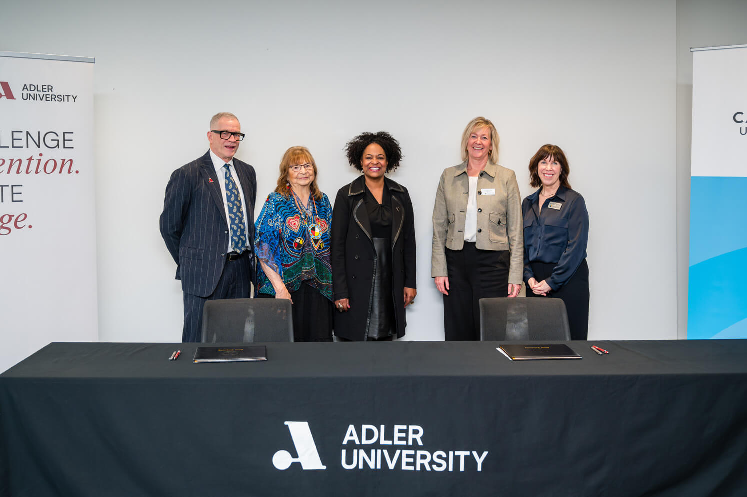 Five people stand behind a table with an Adler University tablecloth, posing for a group photo in front of university banners to celebrate the new Pathway Agreement with Capilano University.