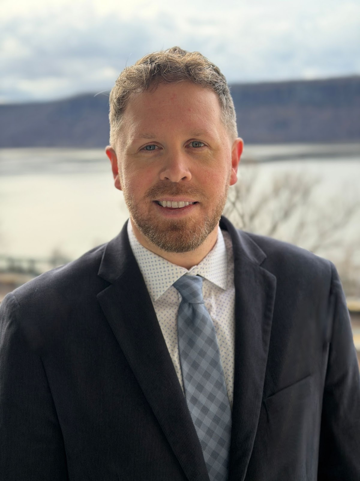 Sean Corlett, dressed in a suit and tie, stands outdoors with a body of water and hills in the background.