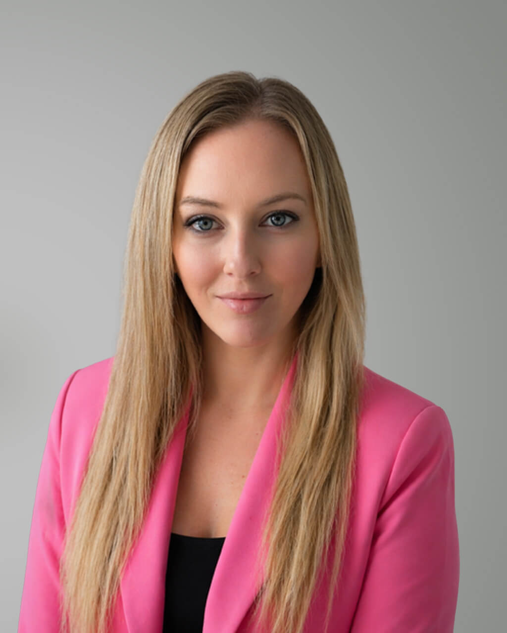 Chelsea Minhas, a woman with long blonde hair, wears a pink blazer over a black top as she poses confidently in front of a plain light grey background.