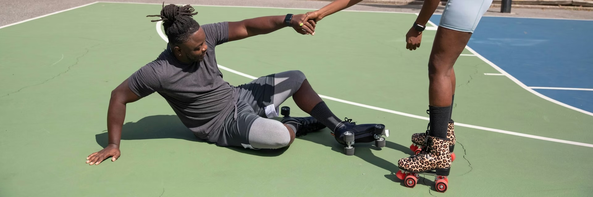 A person wearing roller skates helps another person up from the ground on an outdoor sports court, capturing the spirit of starting a hobby and supporting each other through beginnerhood.