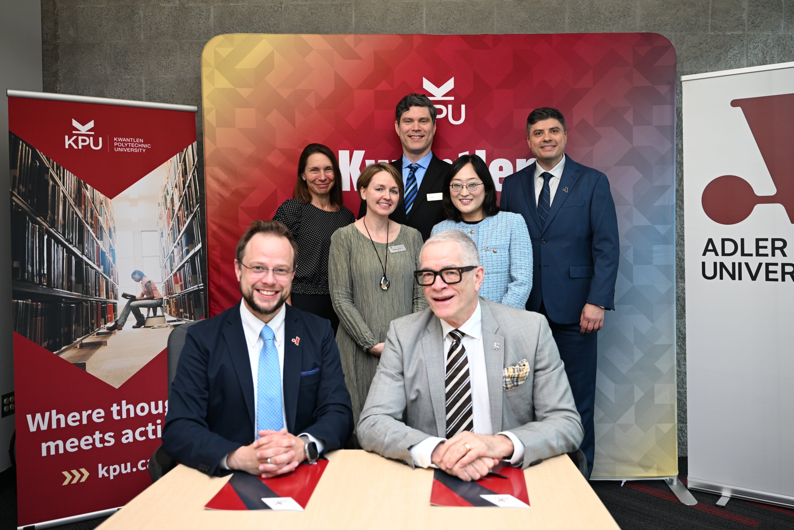 Seven people pose for a group photo at a formal event; two men sit at a table with documents while five others stand behind them, with KPU and Adler University banners in the background.