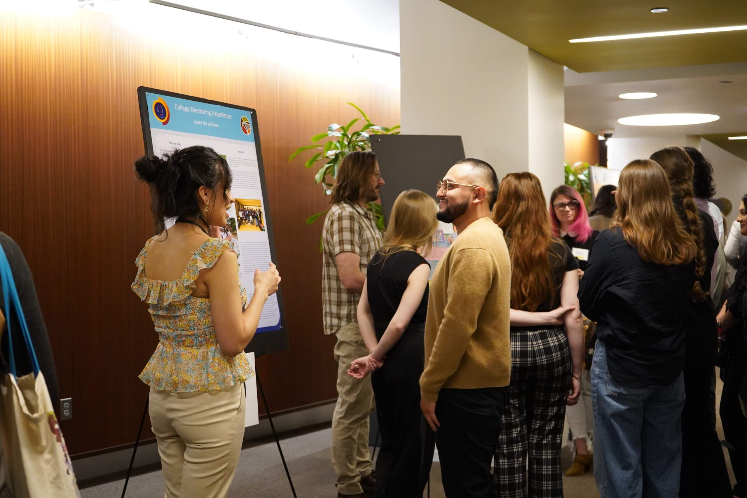 A group of people gather in a hallway during the Practicum Symposium; some read and discuss a poster on an easel while others converse nearby about their community work.