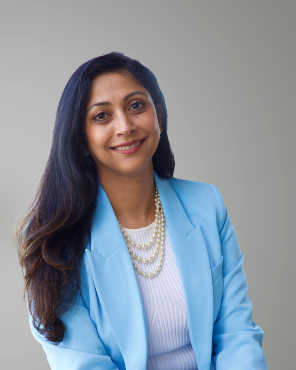 Falguni Shah, une femme aux longs cheveux bruns, porte un blazer bleu, un haut blanc et un collier de perles à plusieurs rangs ; elle sourit à l'appareil photo devant un fond clair et uni.