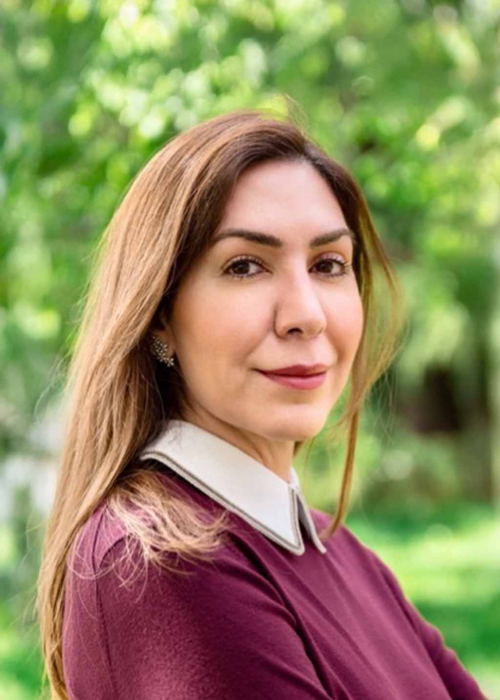 Woman with long brown hair wearing a burgundy top with a white collar, standing outdoors with blurred green foliage in the background.