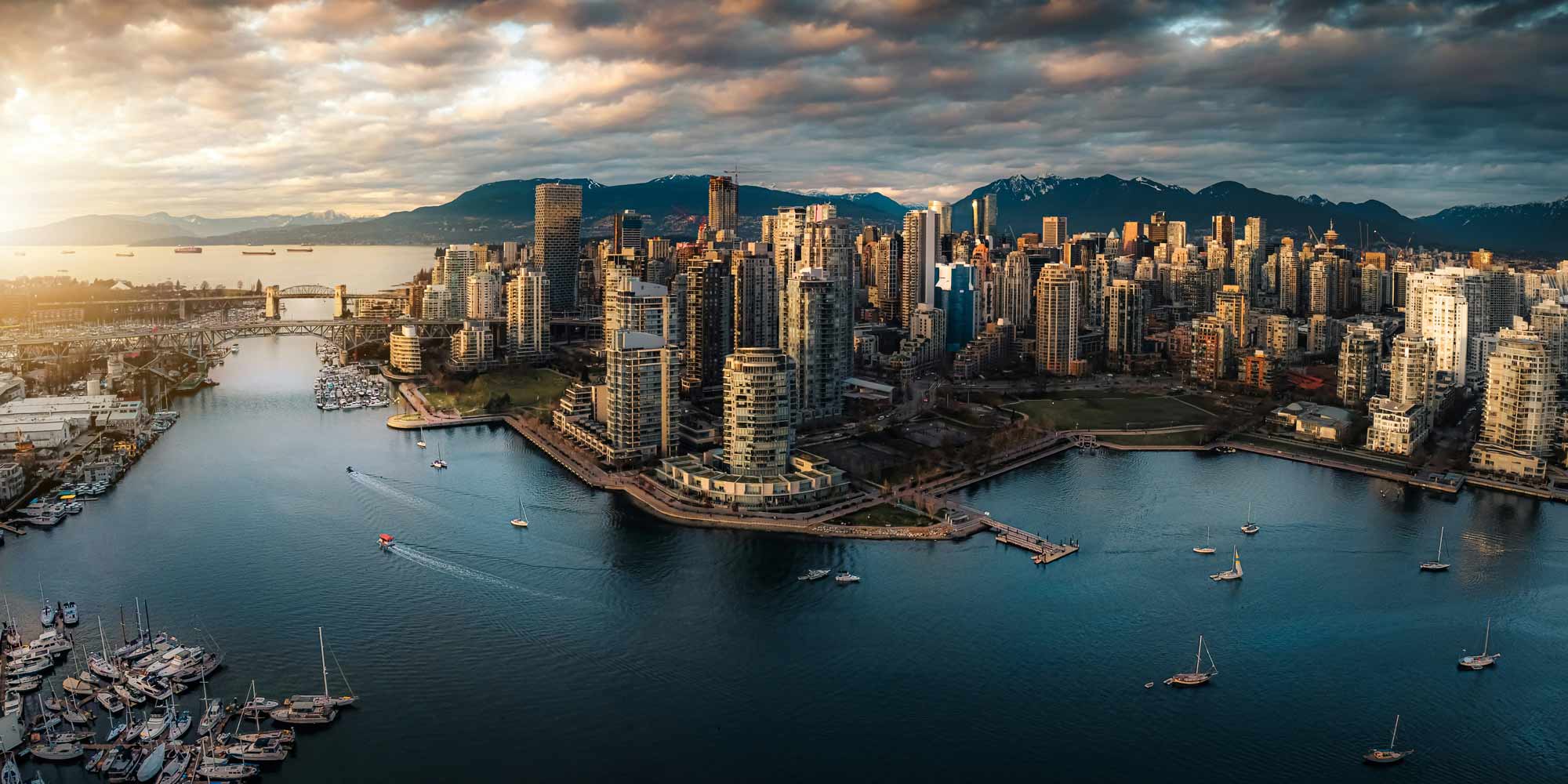 Aerial view of a modern city skyline with high-rise buildings, boats docked in a marina, and mountains in the background under a cloudy sky at sunset—a perfect scene for anyone considering relocating to Vancouver.