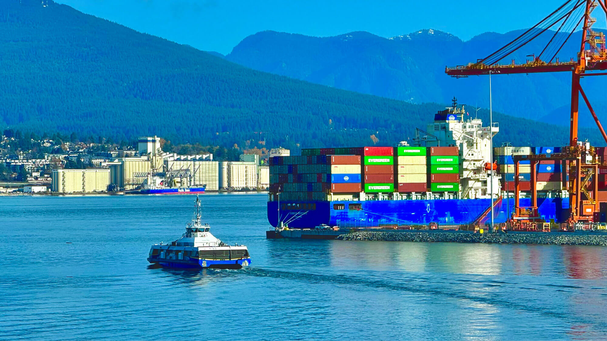 A container ship docked at a port with stacked cargo while a small vessel travels across the water; mountains and industrial buildings are in the background—a familiar scene for those relocating to Vancouver.