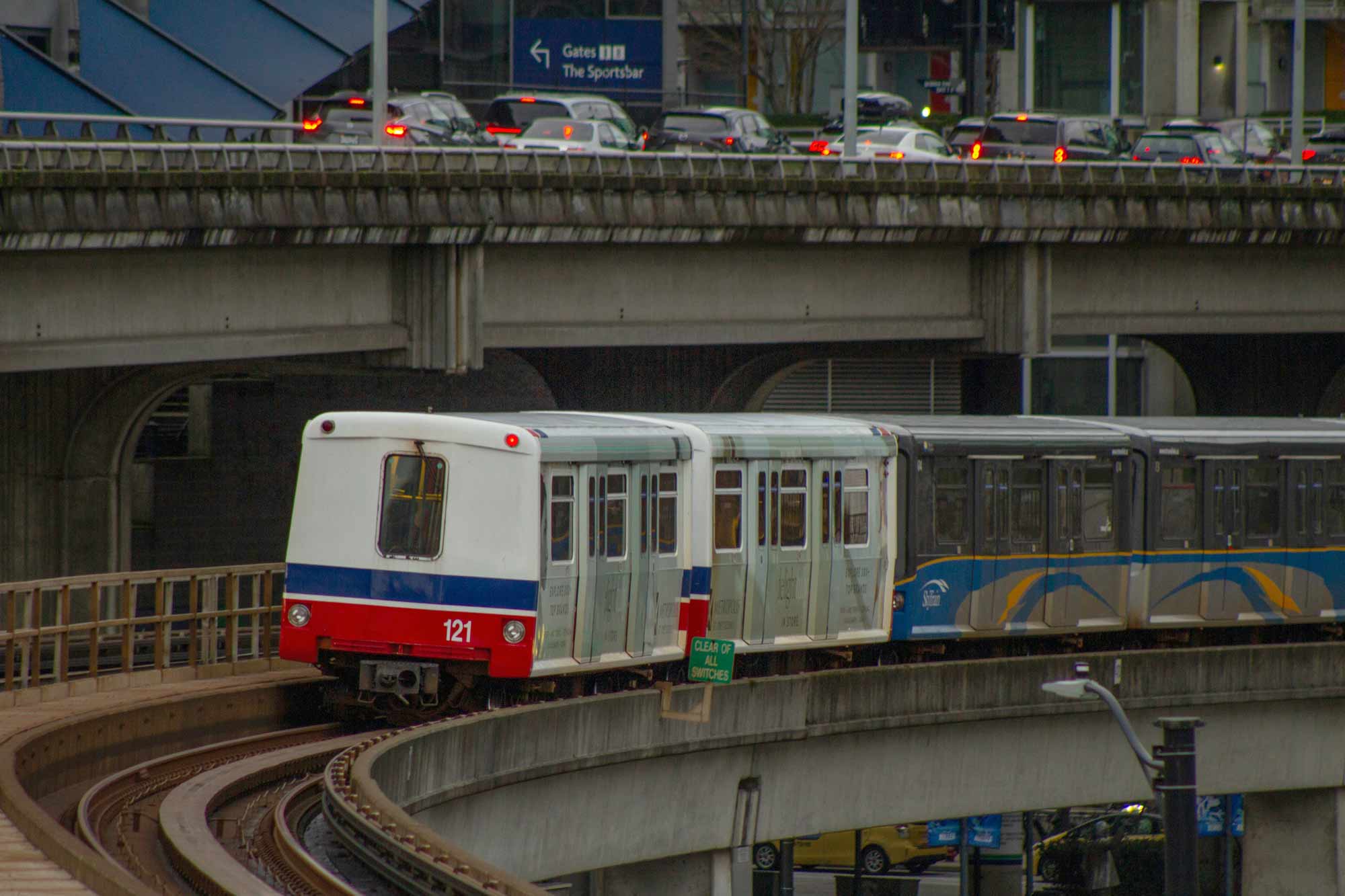 A train with the number 121 travels on an elevated track, with multiple levels of roads and cars visible in the background—a common sight when relocating to Vancouver.