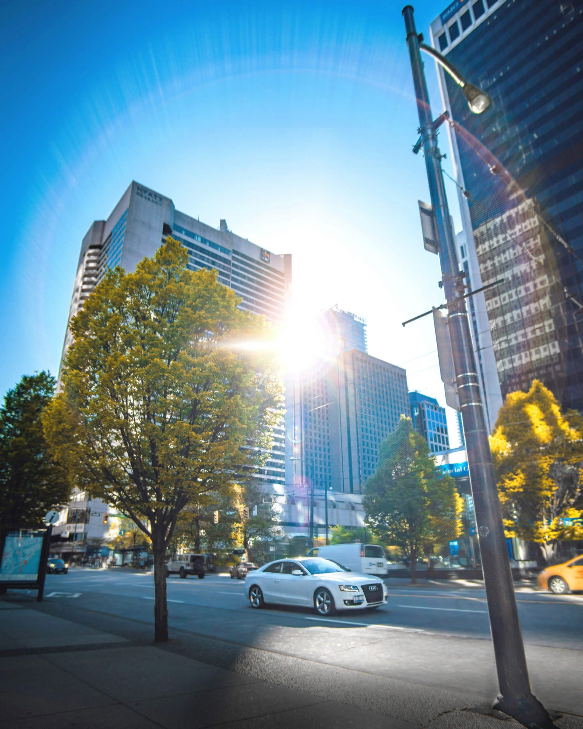 A white car drives down a city street lined with trees and tall buildings under a bright, sunny sky—perfect for anyone relocating to Vancouver and exploring their new city.