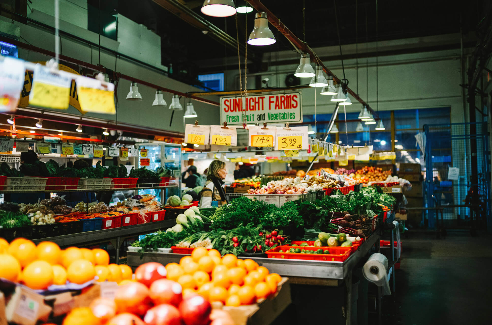 A brightly lit indoor market displays various fresh fruits and vegetables. A person stands behind the produce counter under a "Sunlight Farms" sign, perfect for those relocating to Vancouver and seeking local farm-fresh produce.