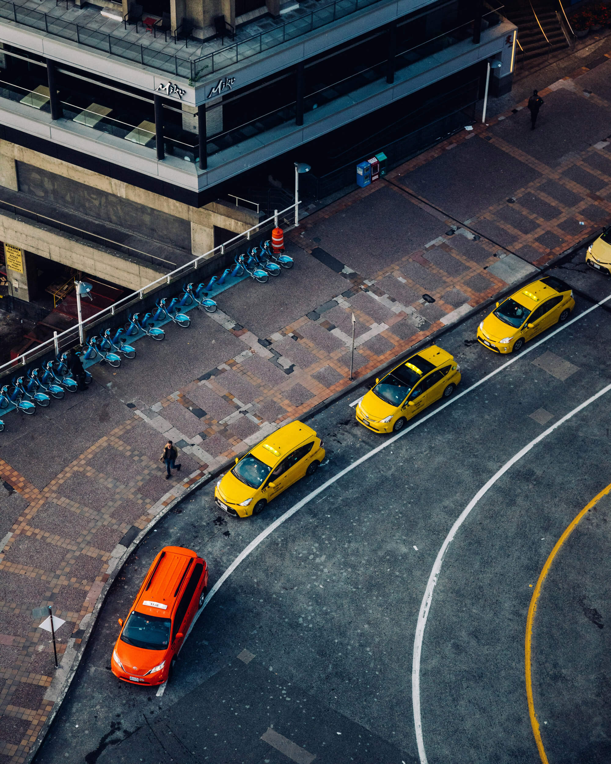 Aerial view of a city street with five yellow taxis and one orange car parked along the curb, near a row of blue bicycles—a typical scene you might spot after relocating to Vancouver.