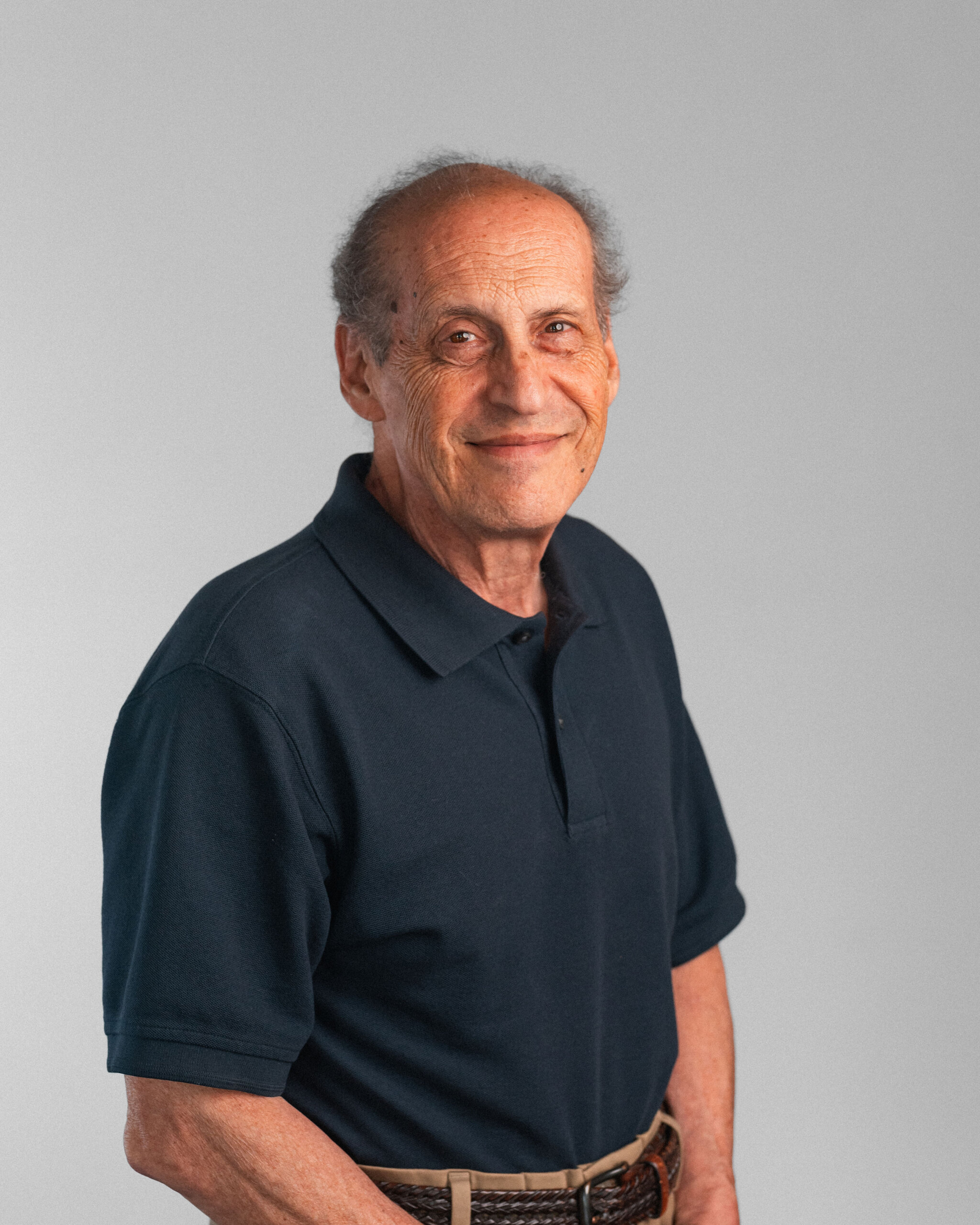 An older man with thinning hair wearing a dark polo shirt stands against a plain light gray background, facing the camera and smiling slightly—an Outstanding International Psychologist recognized by the American Psychological Association.