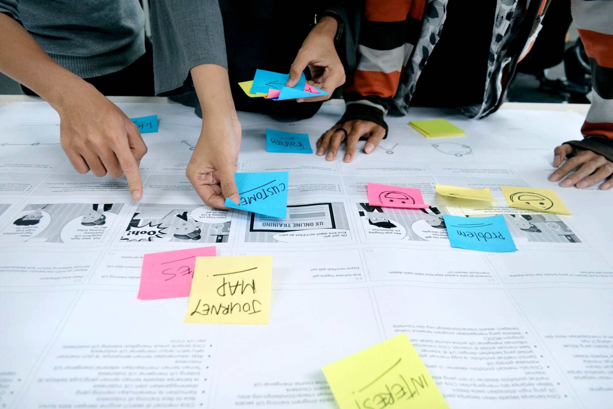 Three people arrange colorful sticky notes on a paper with printed text and comics, collaboratively focusing on planning or brainstorming ideas—a process that reflects principles of shared governance.
