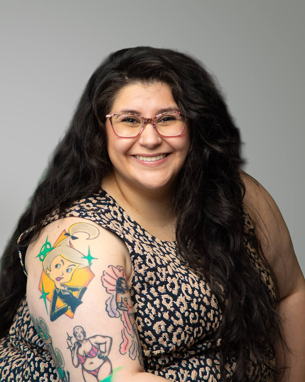 Une femme aux longs cheveux bruns et portant des lunettes sourit à la caméra, dévoilant des tatouages colorés sur le haut de son bras ; elle porte une robe sans manches à motifs. Son style et son assurance reflètent tout le charme naturel de Libby.