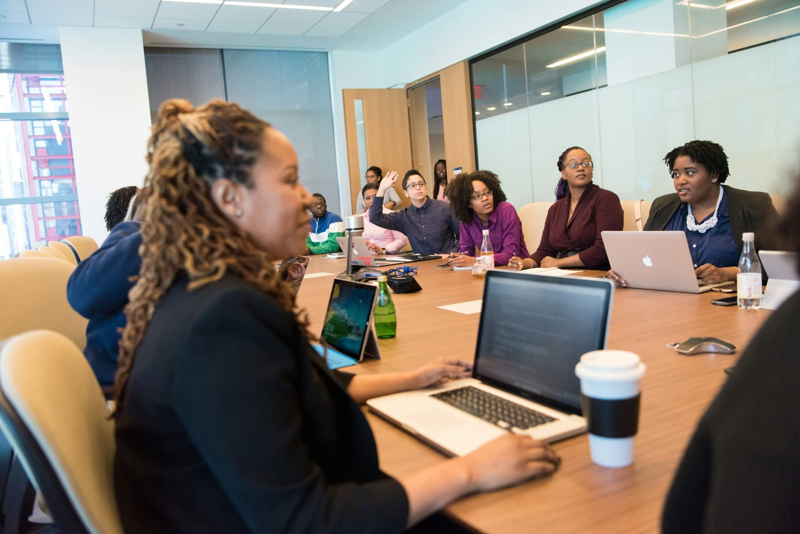 A group of people sit around a conference table with laptops and notebooks, appearing engaged in a meeting or discussion focused on shared governance in a modern office setting.
