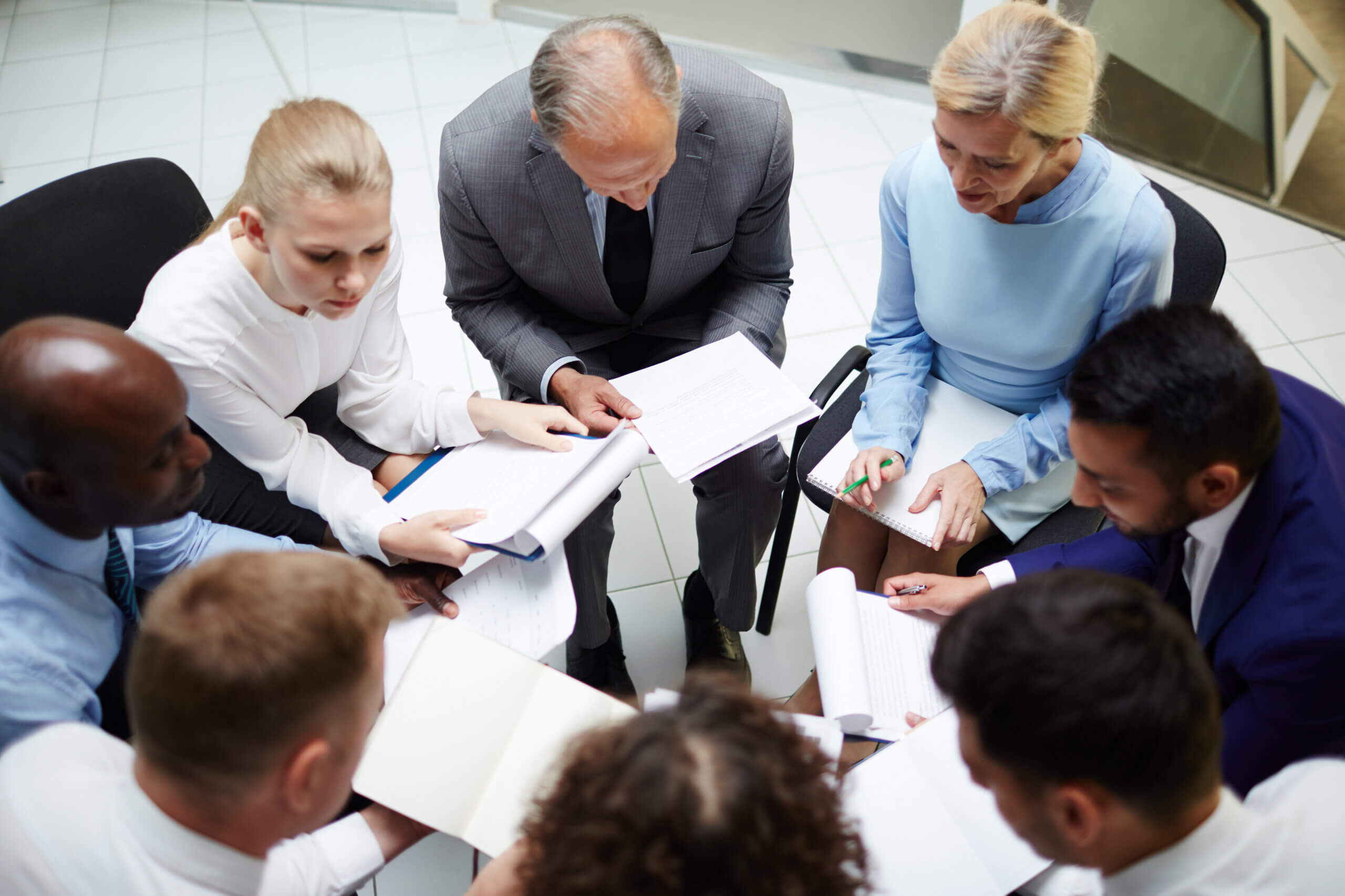 Eight people in business attire sit in a circle, reviewing and discussing documents together in a well-lit office setting, exemplifying shared governance as they collaborate on important decisions.