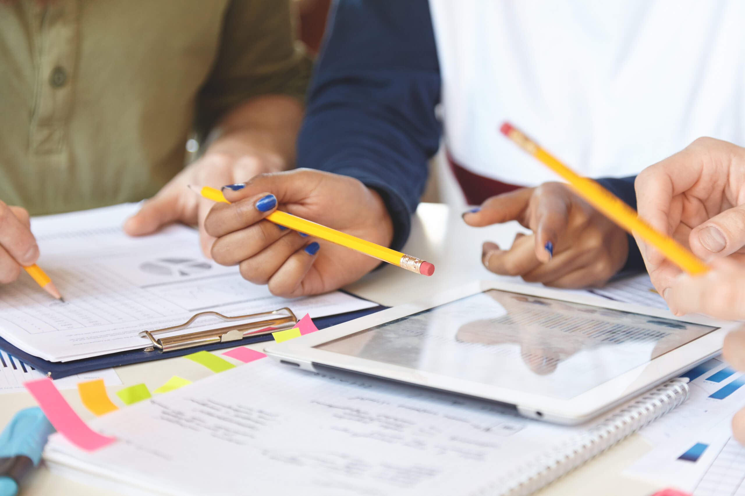 Three people work together at a table with notebooks, papers, pencils, sticky notes, and a tablet, demonstrating shared governance as they collaboratively engage in project planning or group study.