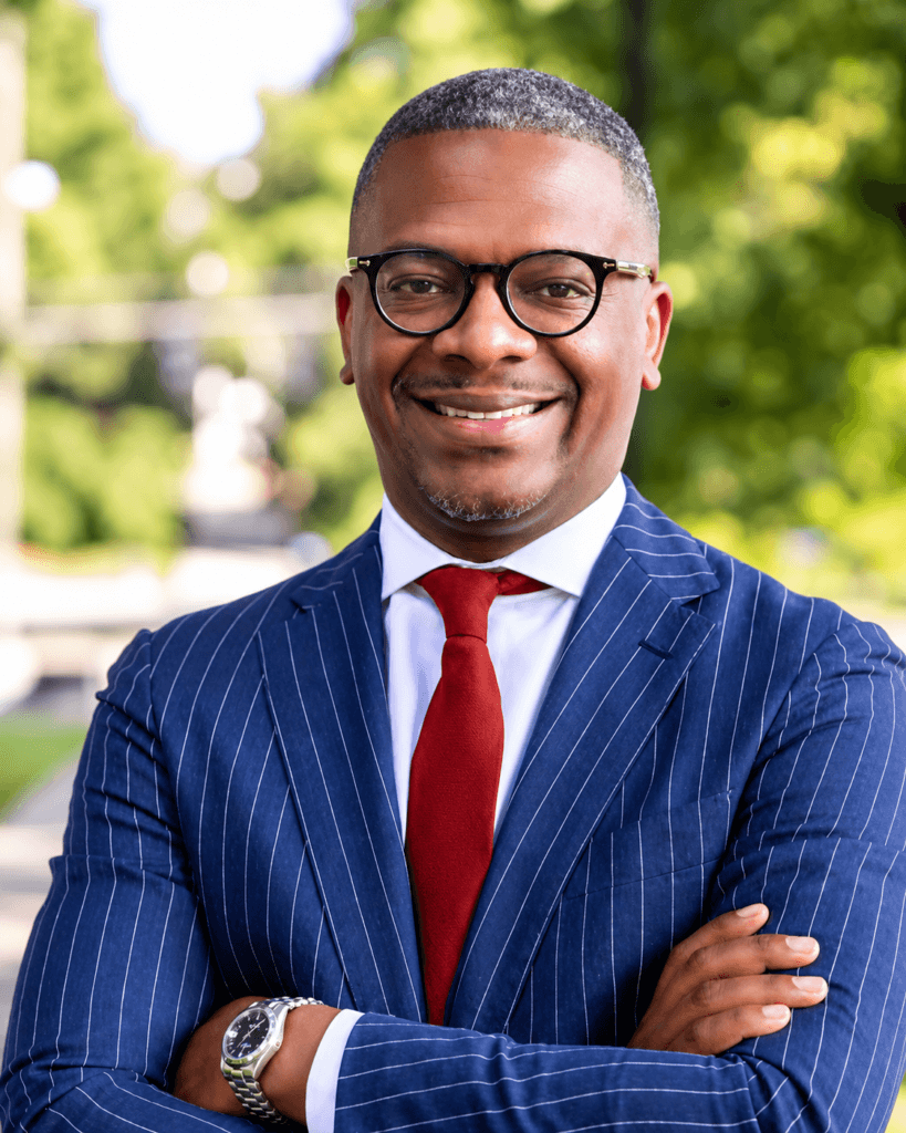 Lewis Williams, a man with glasses, smiles in a navy pinstripe suit, white shirt, and red tie with his arms crossed.